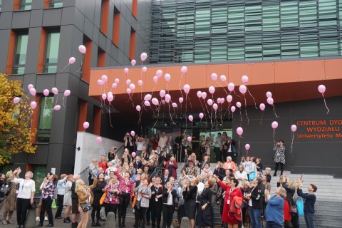 Klikudziesięcioosobowa grupa kobiet, stojąca przed budynkiem, wypuszczająca w niebo różowe balony z helem.