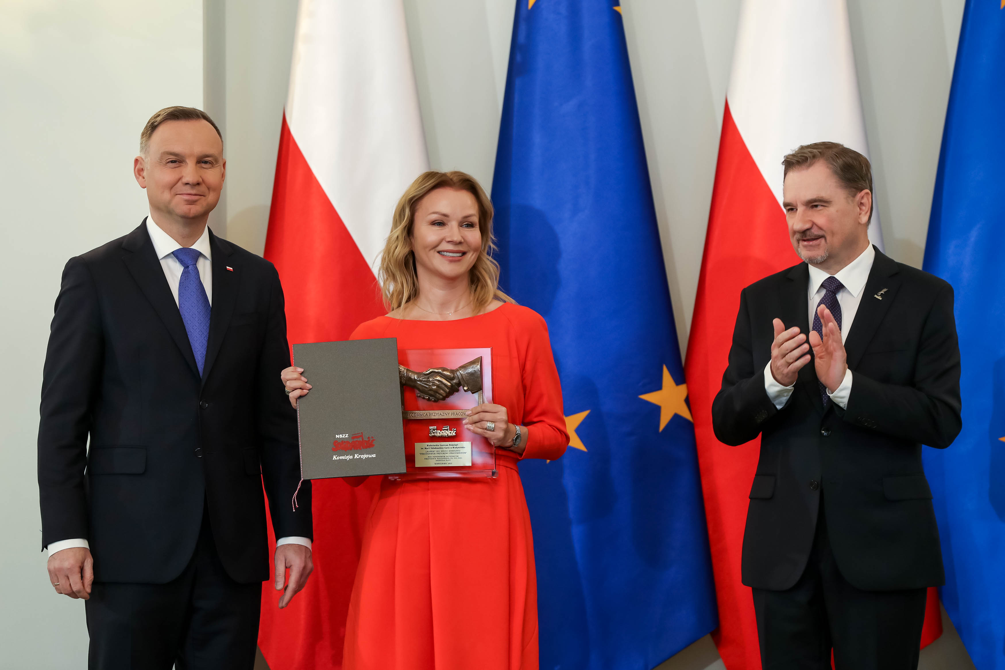 Dwóch mężczyzn w garniturach i kobieta w sukience pozują do zdjęcia na tle flag polskiej i unijnej.